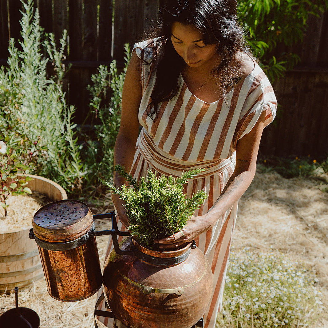 A woman in a cream and rust striped dress stands in a sunny garden next to a rustic wooden barrel, holding a small green plant over a large copper pot while another copper pot hangs beside her.