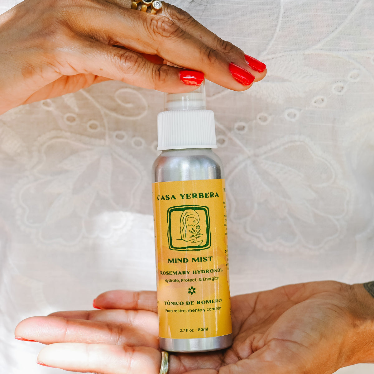 A person holding a small silver bottle labeled Casa Yerbera Mind Mist Rosemary Hydrosol, with both hands visible against a white embroidered garment.