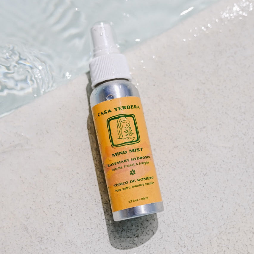 A bottle of Casa Yerberra Mind Mist herbal hydrogel spray with a white spray top and a yellow label, resting on a light stone surface next to water.