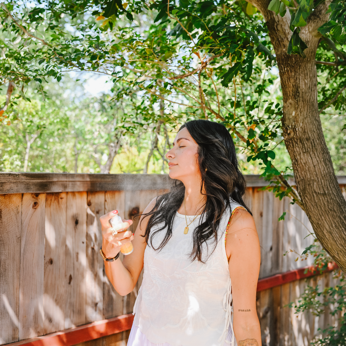 A woman stands outdoors in a backyard garden setting under a tree, wearing a white sleeveless top and white skirt while holding a small bottle.